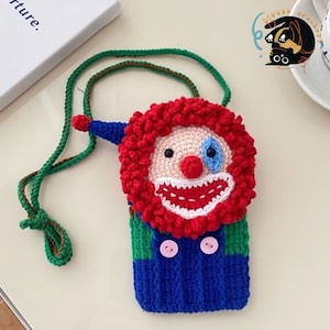 Pu&ograve; includere: Una borsa a mano in crochet a forma di viso da clown con un sorriso rosso e bianco, occhi blu e una parrucca rossa riccia. La borsa &egrave; attaccata a una tracolla verde e marrone.