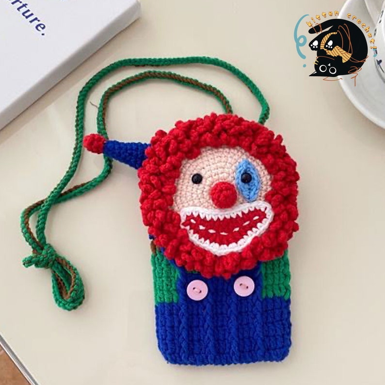 Pu&ograve; includere: Una borsa a mano in crochet a forma di viso da clown con un sorriso rosso e bianco, occhi blu e una parrucca rossa riccia. La borsa &egrave; attaccata a una tracolla verde e marrone.
