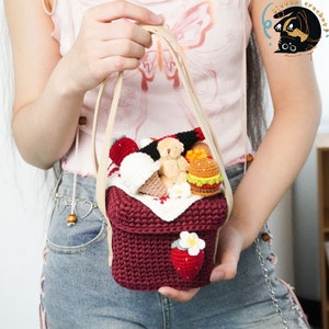 May include: A crocheted picnic basket bag with a red base, white crocheted top, and a strawberry design. The bag is filled with crocheted food items, including a hamburger, ice cream cones, and a teddy bear.