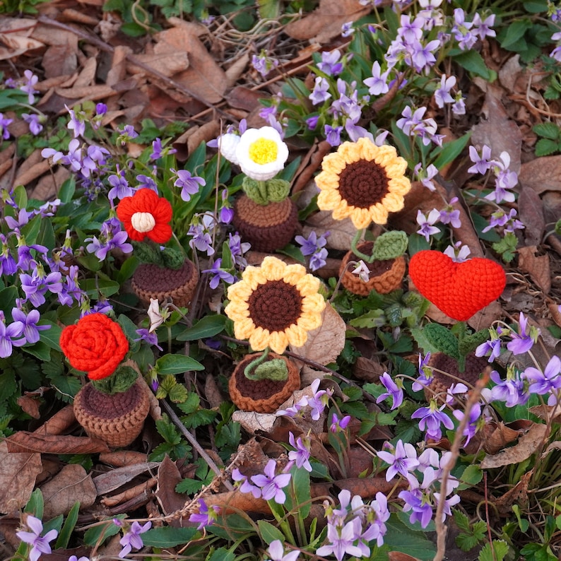 Op de afbeelding: Een verzameling gehaakte bloemen in verschillende kleuren, waaronder rood, wit, geel en oranje, worden gepresenteerd in kleine potten. De bloemen zijn gerangschikt op een bed van groen mos en omringd door paarse bloemen.