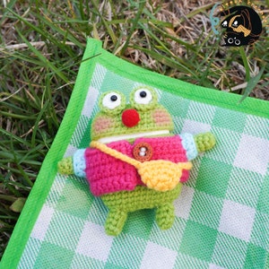 Pode incluir: Um sapo de croch&ecirc; verde vestindo um su&eacute;ter rosa e uma bolsa amarela. O sapo est&aacute; deitado em um cobertor xadrez verde e branco.