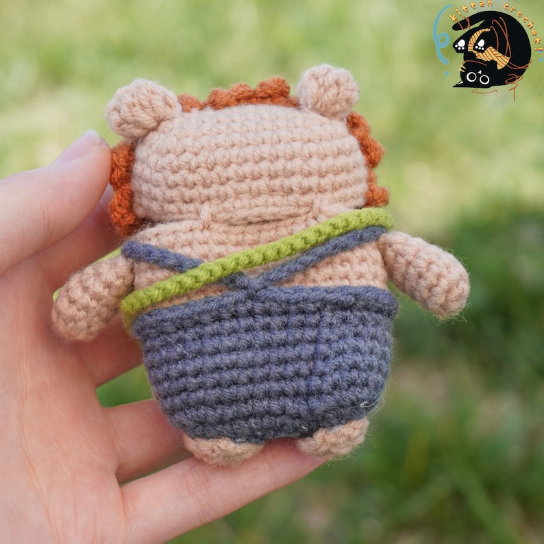 Pode incluir: Um brinquedo de pel&uacute;cia de croch&ecirc; em forma de urso vestindo uma roupa azul e verde. O urso tem pelo marrom e um rosto marrom claro.