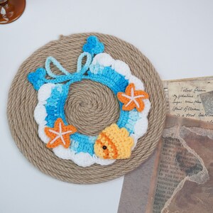 May include: A crocheted wreath in shades of blue, white, and orange, featuring a starfish and a fish design. The wreath is displayed on a brown rope background.