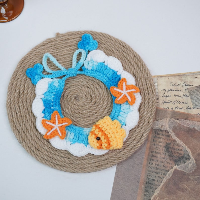 May include: A crocheted wreath in shades of blue, white, and orange, featuring a starfish and a fish design. The wreath is displayed on a brown rope background.