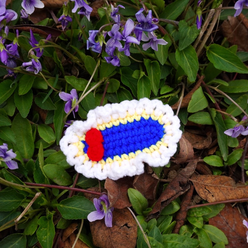 May include: A crocheted blue and white flower with a red center. The flower is lying on a bed of green leaves and brown twigs.