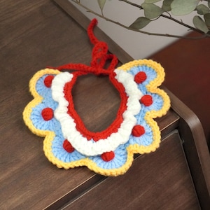 May include: A crocheted pet collar with a red, white, blue, and yellow color scheme. The collar has a scalloped edge and red pom-poms.