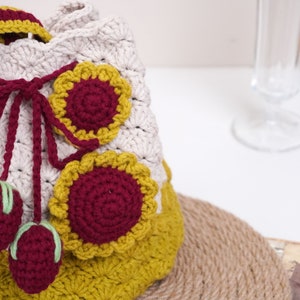 May include: A crocheted bag with a beige base and yellow and burgundy accents. The bag has a drawstring closure and features three burgundy and green pom-poms.