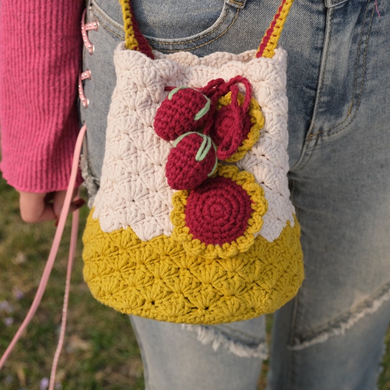 May include: A crocheted bag with a yellow and white pattern. The bag has a drawstring closure and is decorated with red and green crocheted strawberries.