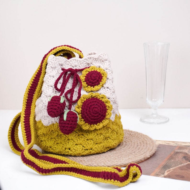 May include: A crocheted bucket bag with a long strap. The bag is made in a beige, mustard yellow, and burgundy color scheme. The bag features a flower design and three small crocheted strawberries.