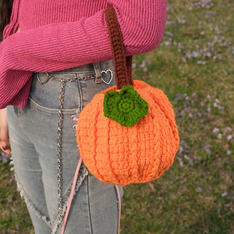 May include: A crocheted pumpkin-shaped bag with a brown handle and a green leaf detail. The bag is worn by a person wearing a pink sweater and blue jeans.