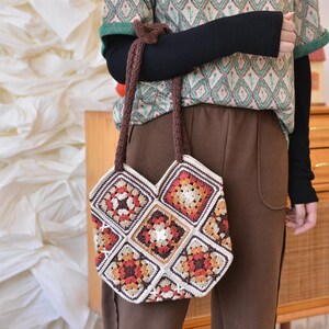 May include: A handmade crochet tote bag featuring a patchwork design with squares in shades of cream, brown, orange, and red. The bag has brown handles and is held by a person wearing a patterned shirt and brown pants.
