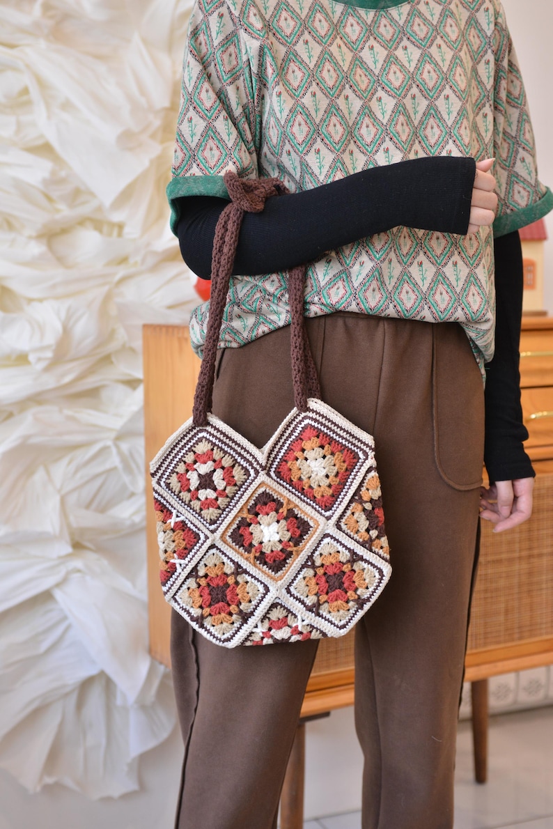 May include: A handmade crochet tote bag featuring a patchwork design with squares in shades of cream, brown, orange, and red. The bag has brown handles and is held by a person wearing a patterned shirt and brown pants.