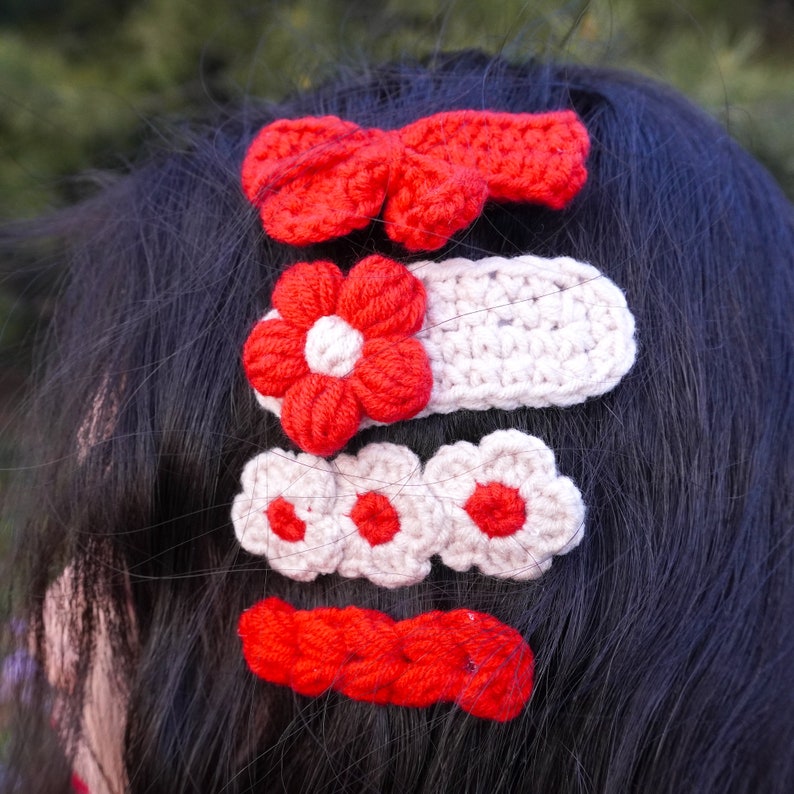 May include: Four crocheted hair clips in red and white. The clips are shaped like a bow, a flower, three small flowers, and a long rectangle.