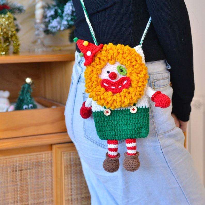 Pu&ograve; includere: Una borsa a forma di clown realizzata a uncinetto con un viso giallo e rosso, pantaloni verdi e gambe a righe bianche e rosse. La borsa viene indossata a tracolla.