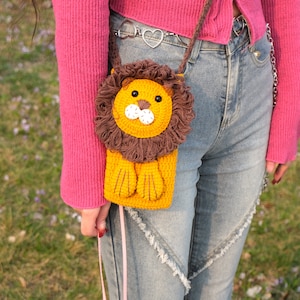 May include: A crocheted lion-shaped purse with brown fur and yellow details. The purse is hanging from a pink strap and is worn by a person wearing blue jeans.