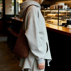 May include: A light gray hoodie with a side pocket and a brown woven shoulder bag. The bag is made of a thick, textured material. The person is standing in a cafe setting.