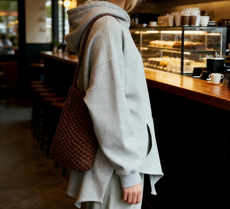 May include: A light gray hoodie with a side pocket and a brown woven shoulder bag. The bag is made of a thick, textured material. The person is standing in a cafe setting.