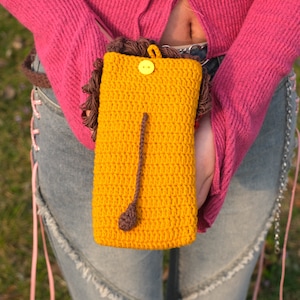 May include: A yellow crocheted phone case with a brown crocheted lion's mane and a yellow button.