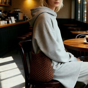 May include: A person wearing a light gray hoodie and matching sweatpants, seated in a cafe. A brown, knitted shoulder bag hangs from the chair. The cafe has wooden tables and chairs, with a coffee cup on the table.