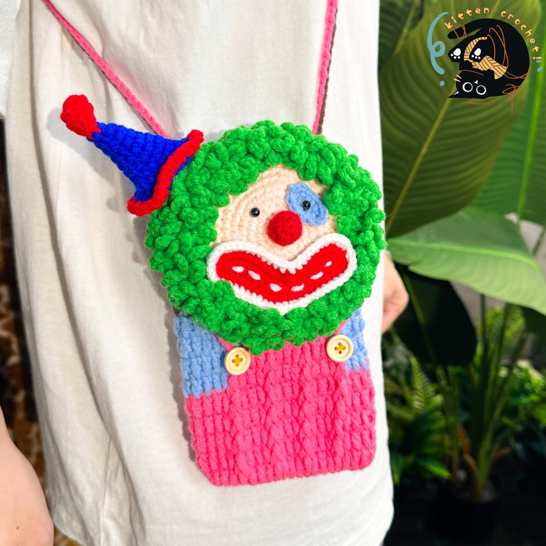 Pu&ograve; includere: Una borsa a tracolla all'uncinetto a forma di clown con un viso verde, una bocca rossa, un occhio blu e una salopette rosa e blu. La borsa ha due bottoni gialli e un cappello rosso e blu.