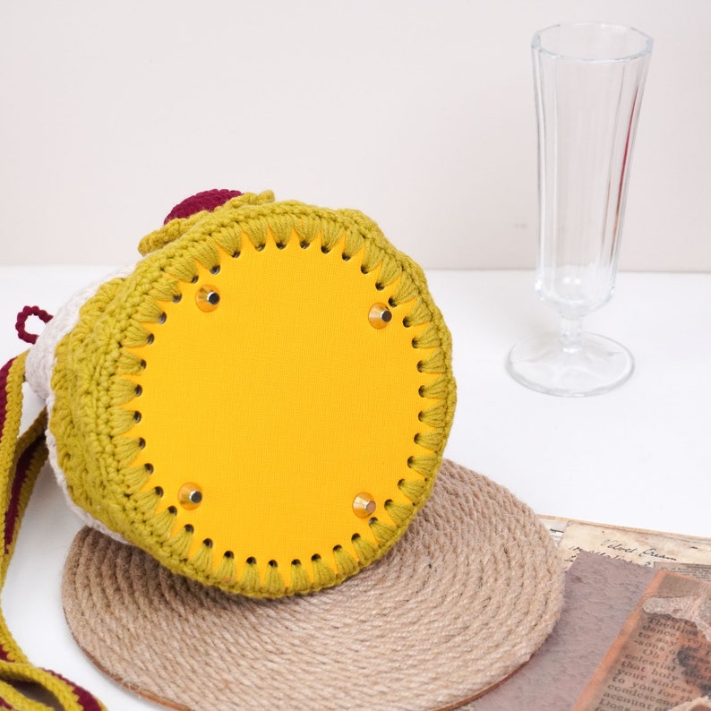 May include: A crocheted bucket bag with a yellow leather bottom and a burgundy strap. The bag has a round shape and is made with a variety of stitches.