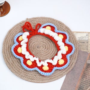 May include: A crocheted collar with a red, white, and blue color scheme. The collar has a scalloped edge and is decorated with small yellow and white pom-poms.