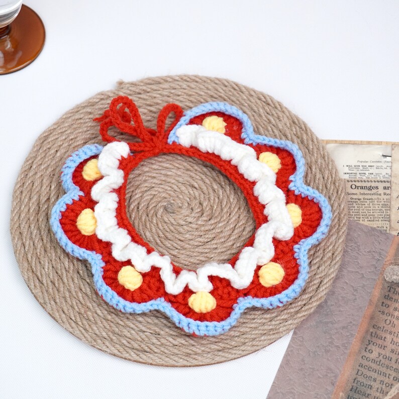 May include: A crocheted collar with a red, white, and blue color scheme. The collar has a scalloped edge and is decorated with small yellow and white pom-poms.