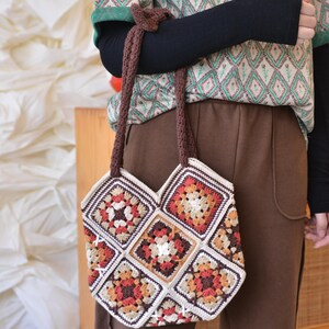 May include: A handmade crochet tote bag featuring a patchwork design with squares in shades of cream, brown, orange, and red. The bag has a brown strap and is held by a person. The bag's design is reminiscent of a granny square pattern.
