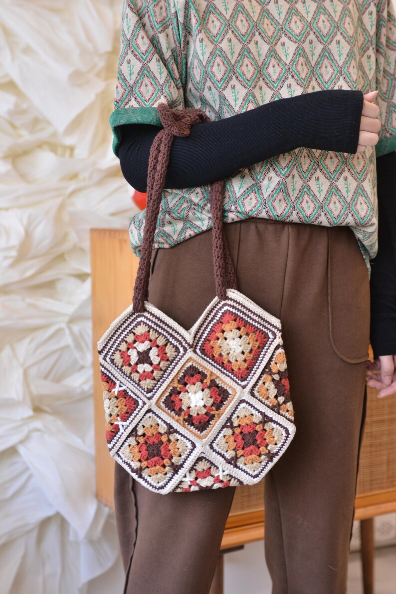 May include: A handmade crochet tote bag featuring a patchwork design with squares in shades of cream, brown, orange, and red. The bag has a brown strap and is held by a person. The bag's design is reminiscent of a granny square pattern.