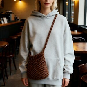 May include: A brown, crocheted shoulder bag with a long strap. The bag is a small, bucket-style design. The strap is a braided style. The person in the image is wearing a gray hoodie.