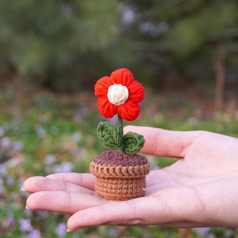 Op de afbeelding: Een klein gehaakt bloemetje in een pot. De bloem is rood met een wit hartje en groene blaadjes. De pot is bruin.