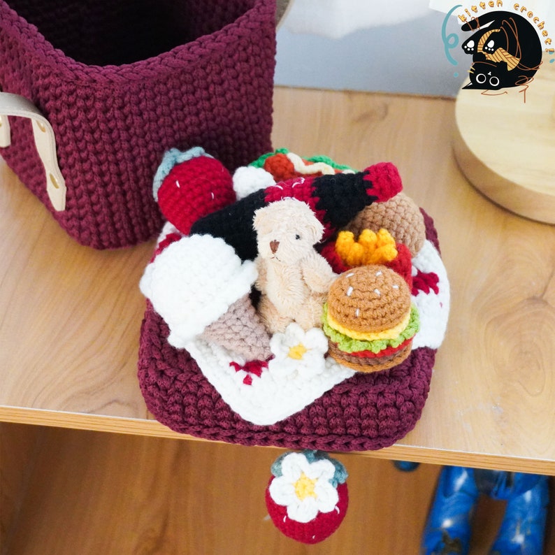 May include: A crocheted picnic basket filled with miniature crocheted food items, including a hamburger, chips, a bottle of ketchup, a teddy bear, and a strawberry. The basket is a deep burgundy colour and is sitting on a wooden surface.