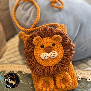 May include: A crocheted lion-shaped purse with a brown mane, orange face, and white nose and eyes. The purse has a brown strap and is made with a soft, textured yarn.