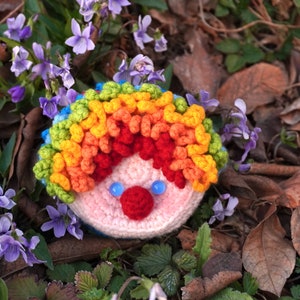 May include: A crocheted clown doll with a rainbow-colored wig, blue eyes, and a red nose. The doll is sitting on a bed of purple flowers and brown leaves.