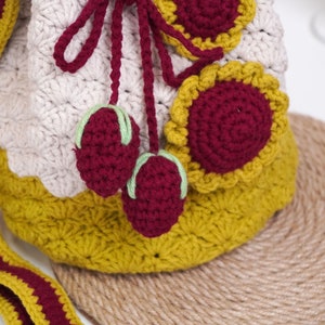 May include: A crocheted drawstring bag with a beige, yellow, and burgundy color scheme. The bag has a flower design and two burgundy cherries attached to the drawstring.