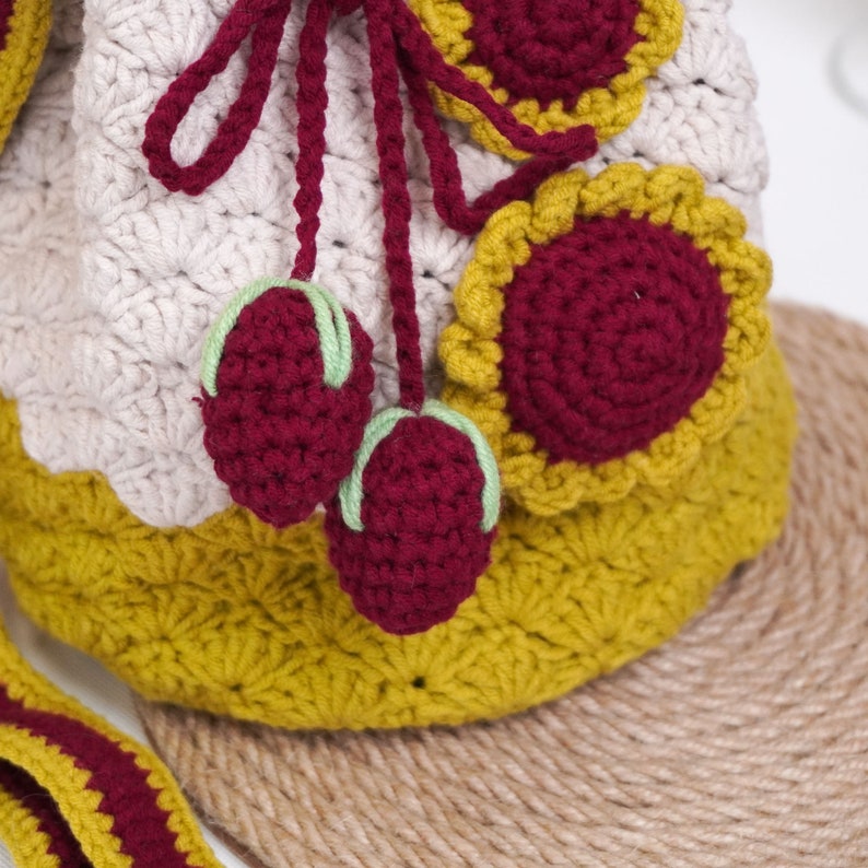 May include: A crocheted drawstring bag with a beige, yellow, and burgundy color scheme. The bag has a flower design and two burgundy cherries attached to the drawstring.
