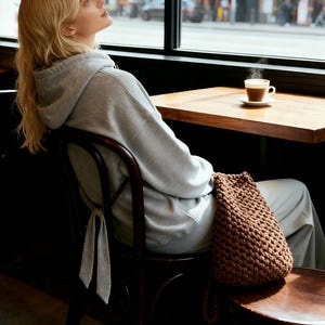 May include: A woman wearing a gray hoodie and sweatpants sits in a cafe. A brown, knitted drawstring bag rests on the chair. A cup of coffee sits on a wooden table near a window.