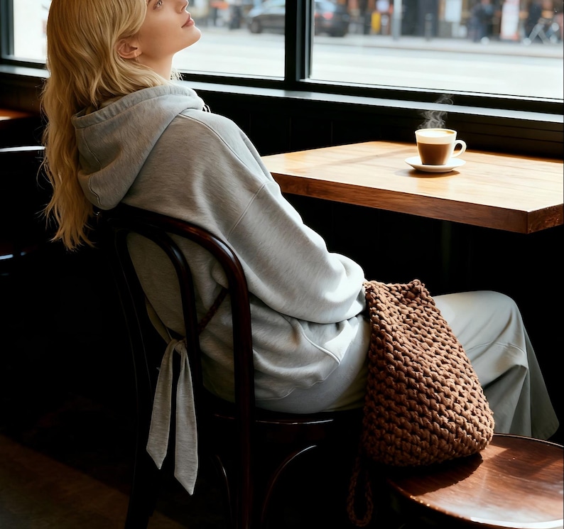 May include: A woman wearing a gray hoodie and sweatpants sits in a cafe. A brown, knitted drawstring bag rests on the chair. A cup of coffee sits on a wooden table near a window.
