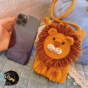 May include: A crocheted lion-shaped purse with a brown strap. The purse is made of orange and brown yarn and has a white face with black eyes.