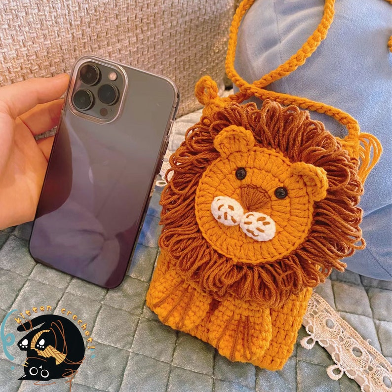 May include: A crocheted lion-shaped purse with a brown strap. The purse is made of orange and brown yarn and has a white face with black eyes.
