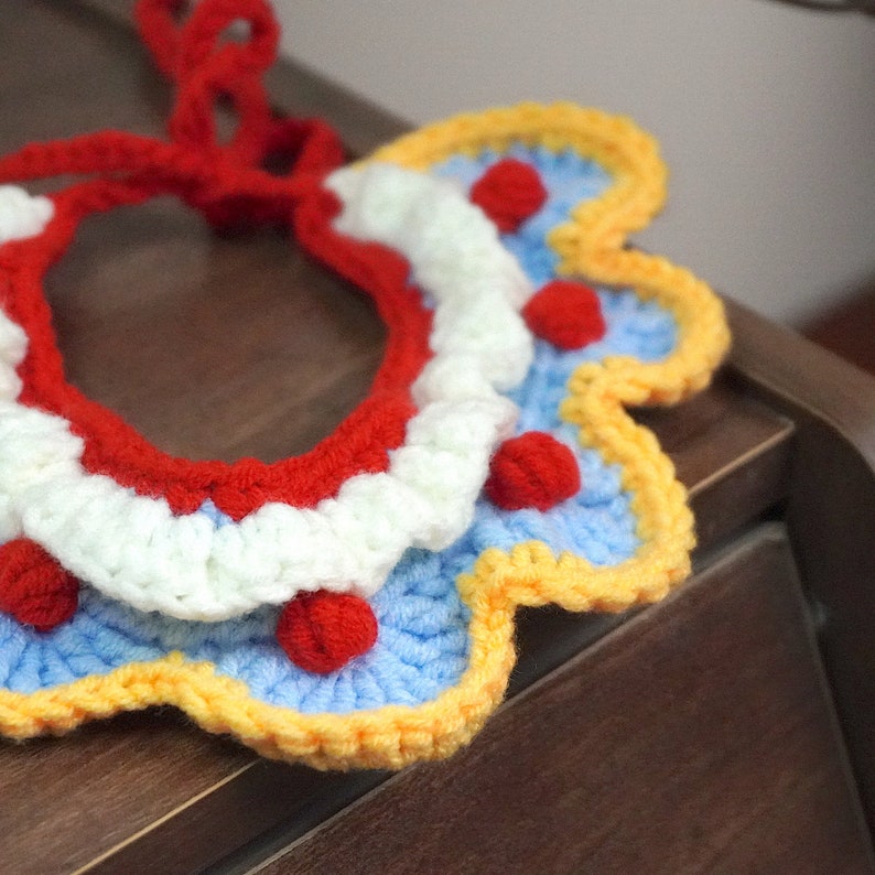May include: A crocheted pet collar with a red, white, blue, and yellow color scheme. The collar has a scalloped edge and features small red pom-poms.