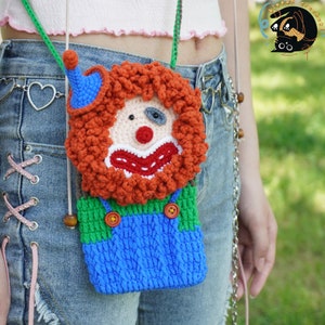 Pu&ograve; includere: Una borsa a mano a forma di clown realizzata a uncinetto con capelli rossi e arancioni, salopette blu e camicia verde. La borsa ha una perla di legno marrone sulla tracolla e viene indossata sulla spalla di una persona.