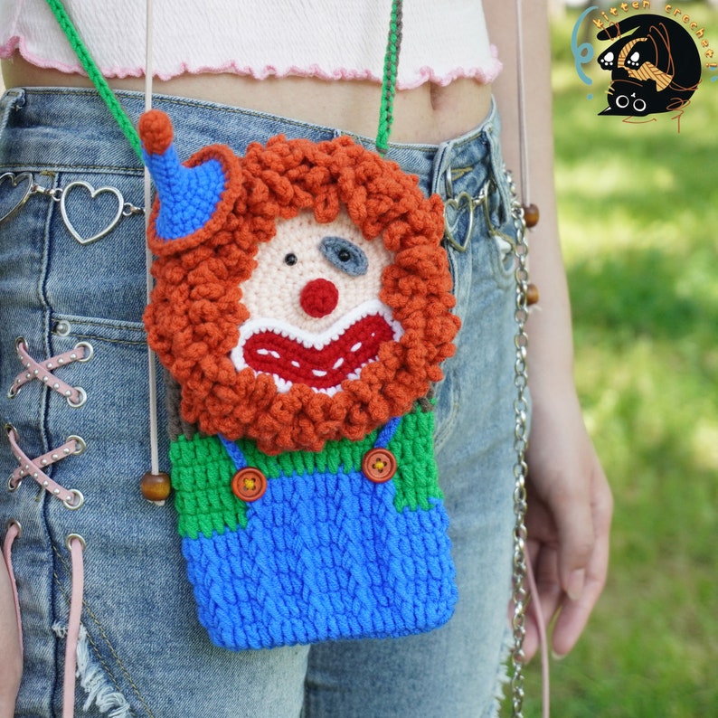 Pu&ograve; includere: Una borsa a mano a forma di clown realizzata a uncinetto con capelli rossi e arancioni, salopette blu e camicia verde. La borsa ha una perla di legno marrone sulla tracolla e viene indossata sulla spalla di una persona.