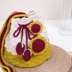 May include: A crocheted bucket bag with a mustard yellow and cream color scheme. The bag has a red and yellow flower design and a red cherry design. The bag has a long strap.