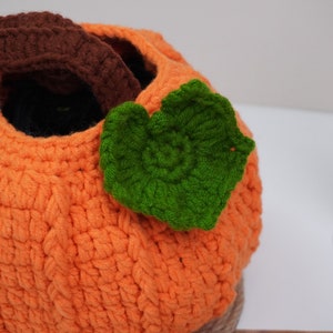 May include: A crocheted pumpkin-shaped basket with a green leaf on top. The basket is made of orange yarn and has a brown yarn loop at the top.
