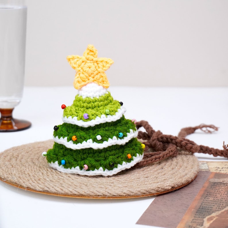 Puede incluir: Un adorno de &aacute;rbol de Navidad tejido a crochet con una estrella amarilla en la parte superior. El &aacute;rbol es verde y blanco y tiene cuentas de colores para decoraci&oacute;n. El adorno est&aacute; sentado en una estera tejida marr&oacute;n.