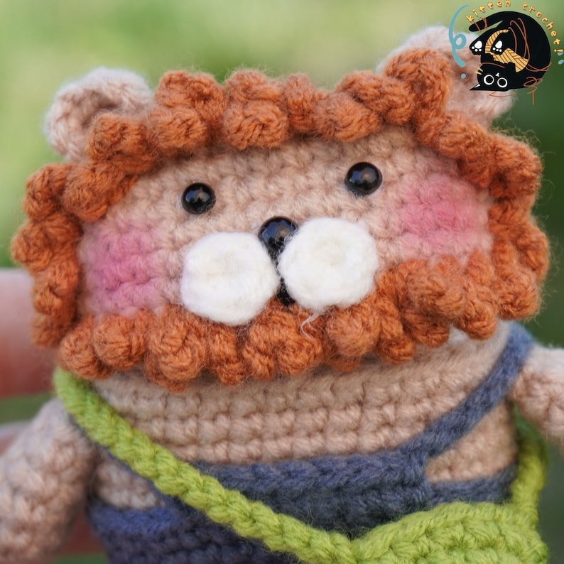 Pode incluir: Um brinquedo de le&atilde;o de croch&ecirc; com uma juba marrom, um corpo bege e uma roupa azul e verde. O le&atilde;o tem um nariz branco e bigodes brancos.