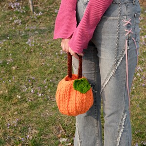 May include: A crocheted pumpkin-shaped bag with a brown handle and a green leaf detail. The bag is being held by a person wearing a pink sweater and blue jeans.