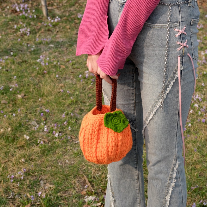 May include: A crocheted pumpkin-shaped bag with a brown handle and a green leaf detail. The bag is being held by a person wearing a pink sweater and blue jeans.