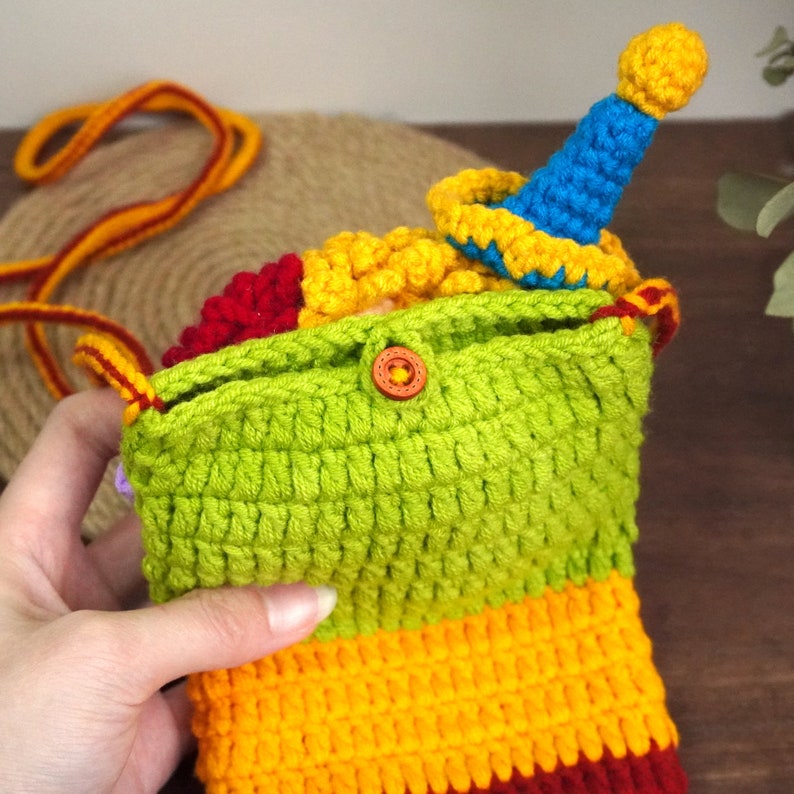 Pu&ograve; includere: Una borsa all'uncinetto colorata con una patta verde e un fondo a righe rosso, giallo e arancione. La borsa contiene una piccola bambola di clown all'uncinetto con un cappello blu.